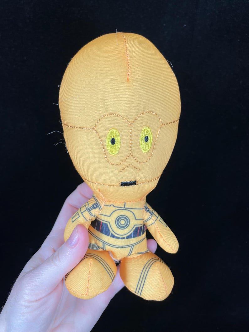 Op de afbeelding: Een pluche speelgoed van het Star Wars-personage C-3PO. De pop is voornamelijk geel met zwarte en grijze details. Het heeft geborduurde kenmerken, waaronder gele ogen en een zwarte mond. Het speelgoed wordt door een hand vastgehouden.