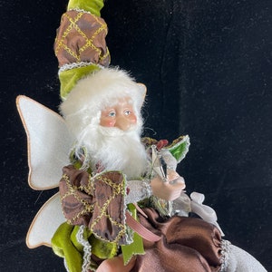 May include: A whimsical fairy figurine with a white beard and wings, wearing a green and brown outfit with gold accents. The fairy has a pointed hat and is holding a pen. The figurine is a decorative item.