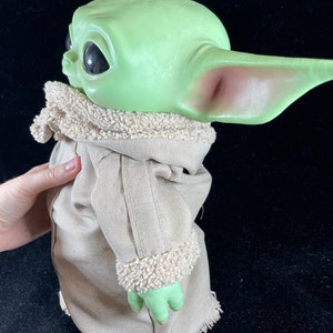 May include: A green plush toy with large ears and big eyes, wearing a brown coat with a white fur trim. The toy is a popular character from the Star Wars franchise.