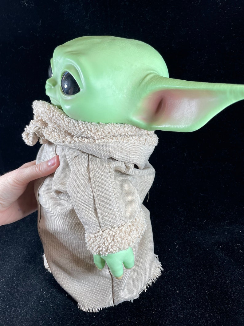 May include: A green plush toy with large ears and big eyes, wearing a brown coat with a white fur trim. The toy is a popular character from the Star Wars franchise.