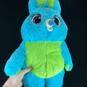 May include: A plush toy bunny in shades of blue and green. The bunny has large, expressive eyes with purple accents, and a small, white nose. The ears are tipped with green, and the belly is a vibrant green color. The toy is soft and cuddly.