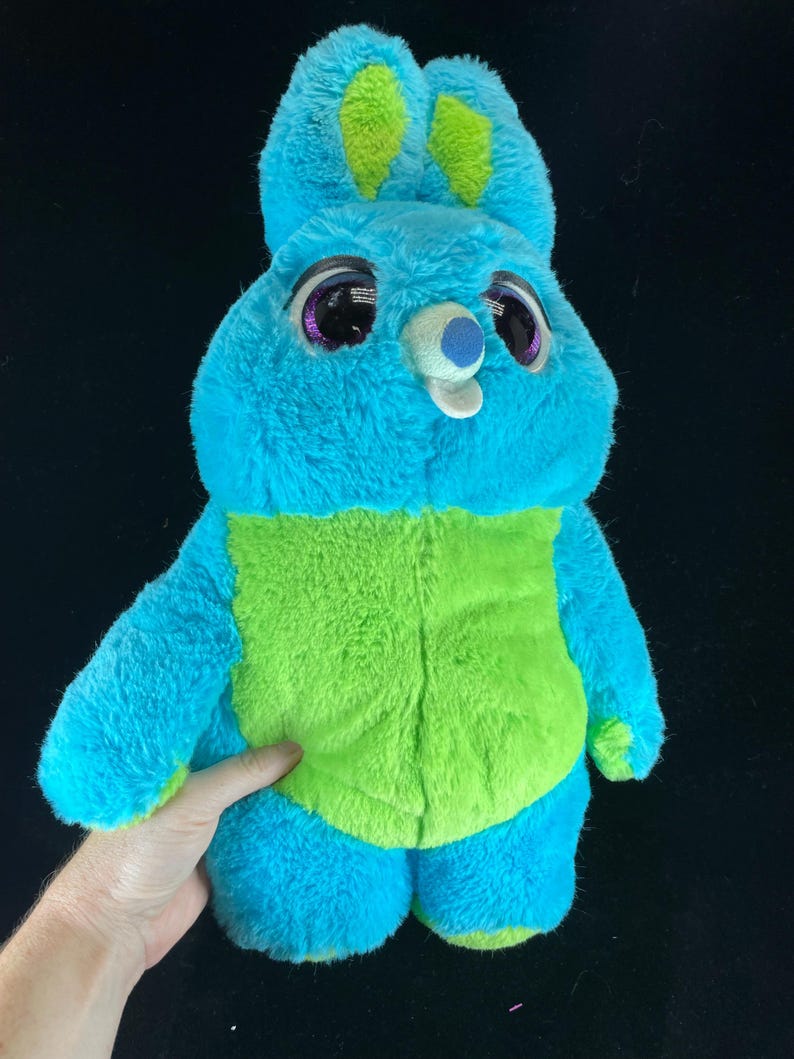 May include: A plush toy bunny in shades of blue and green. The bunny has large, expressive eyes with purple accents, and a small, white nose. The ears are tipped with green, and the belly is a vibrant green color. The toy is soft and cuddly.