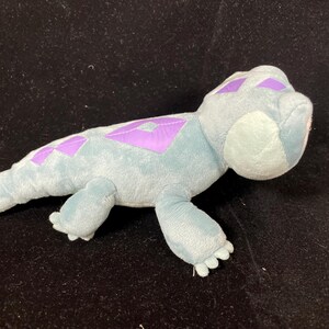 May include: A plush toy lizard in shades of blue and purple. The soft toy features a light blue body with purple diamond-shaped markings along its back. The lizard has small, detailed feet and a rounded head.