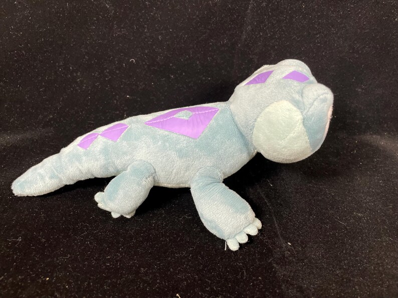 May include: A plush toy lizard in shades of blue and purple. The soft toy features a light blue body with purple diamond-shaped markings along its back. The lizard has small, detailed feet and a rounded head.
