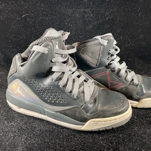 May include: A pair of black high-top sneakers with gray laces and a silver Jumpman logo on the side. The shoes have a white sole and a red stitching detail on the back.