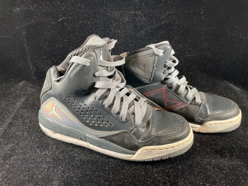 May include: A pair of black high-top sneakers with gray laces and a silver Jumpman logo on the side. The shoes have a white sole and a red stitching detail on the back.