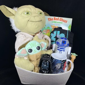 Op de afbeelding: Een grijze mand gevuld met Star Wars-items. Inclusief een groot Yoda-knuffel, een Baby Yoda-knuffel, een boek getiteld "The Red Ghost", een zwart t-shirt, een R2-D2-figuur, een mok en een Darth Vader-figuur.