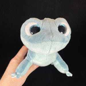 May include: A plush toy frog in shades of blue, with large, round eyes and a soft, velvety texture. The frog has a light blue body and darker blue accents, with a friendly, cartoonish appearance.
