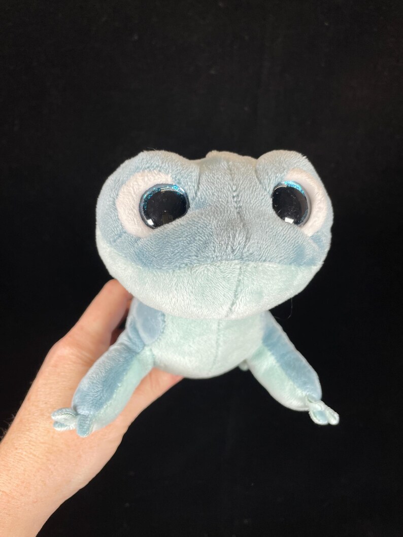 May include: A plush toy frog in shades of blue, with large, round eyes and a soft, velvety texture. The frog has a light blue body and darker blue accents, with a friendly, cartoonish appearance.