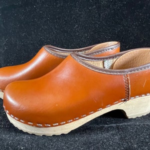 May include: A pair of brown leather clogs with wooden soles. The clogs have a simple design and are a classic style.