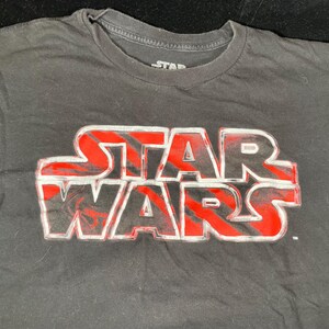 Op de afbeelding: Zwart t-shirt met het Star Wars-logo in rood en wit. Het logo staat prominent op de borst. Het shirt heeft een ronde hals en korte mouwen. Het Star Wars-logo is een klassiek ontwerp.