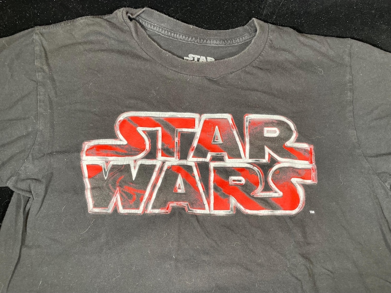 Op de afbeelding: Zwart t-shirt met het Star Wars-logo in rood en wit. Het logo staat prominent op de borst. Het shirt heeft een ronde hals en korte mouwen. Het Star Wars-logo is een klassiek ontwerp.