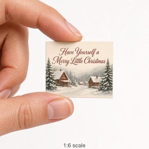 Może przedstawiać: Kwadratowa miniaturowa karta przedstawiająca zimowy krajobraz z czerwonymi domami, ośnieżonymi sosnami i napisem Have Yourself a Merry Little Christmas czerwoną czcionką. Karta jest oznaczona jako skala 1:6 rzeczywisty rozmiar.
