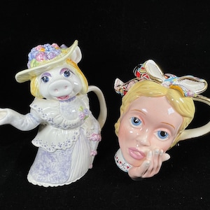 Puede incluir: Dos teteras de cerámica. Una tiene forma de cerdo con sombrero y vestido, la otra es una cara de mujer con un lazo. Ambas tienen asas doradas y están sobre un fondo negro.