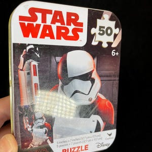 Op de afbeelding: Een Star Wars-puzzel in een metalen doos. De doos toont een Stormtrooper-afbeelding en het Star Wars-logo in rood en wit. De puzzel is 12,7 cm x 17,7 cm groot en bevat 50 stukjes. Aanbevolen voor kinderen vanaf 6 jaar.
