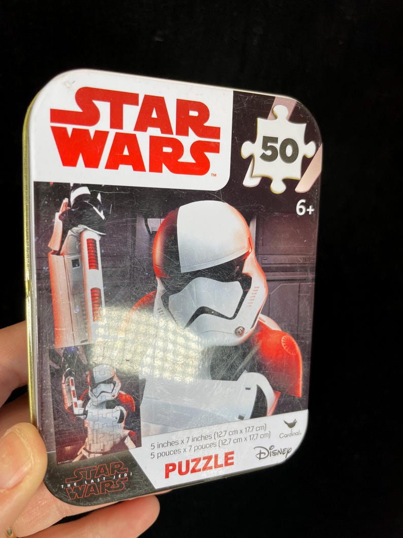 Op de afbeelding: Een Star Wars-puzzel in een metalen doos. De doos toont een Stormtrooper-afbeelding en het Star Wars-logo in rood en wit. De puzzel is 12,7 cm x 17,7 cm groot en bevat 50 stukjes. Aanbevolen voor kinderen vanaf 6 jaar.