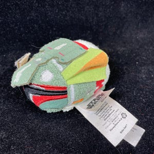 May include: A small, plush Boba Fett character from Star Wars. The toy features a green helmet with orange and yellow accents, red details, and a white body. A white tag with text is attached.