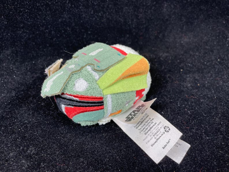 May include: A small, plush Boba Fett character from Star Wars. The toy features a green helmet with orange and yellow accents, red details, and a white body. A white tag with text is attached.