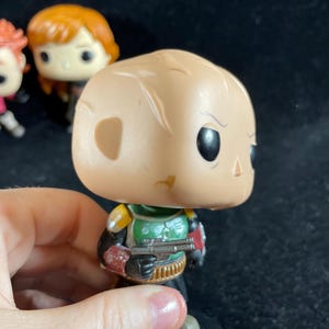 May include: A Funko Pop! vinyl figure of Boba Fett from Star Wars. The figure has a tan head, green and yellow armour, and a blaster. Other figures are visible in the background.