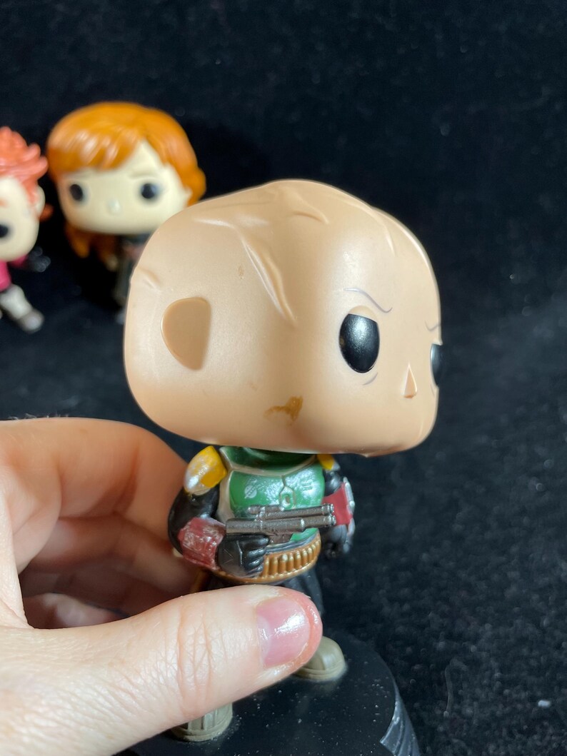 May include: A Funko Pop! vinyl figure of Boba Fett from Star Wars. The figure has a tan head, green and yellow armour, and a blaster. Other figures are visible in the background.