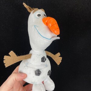 May include: A plush snowman toy with a carrot nose, brown twig arms, and a small brown twig on top of its head. The snowman has black glitter buttons and snowflake accents. The toy is white with blue embroidered eyes and a blue smile.