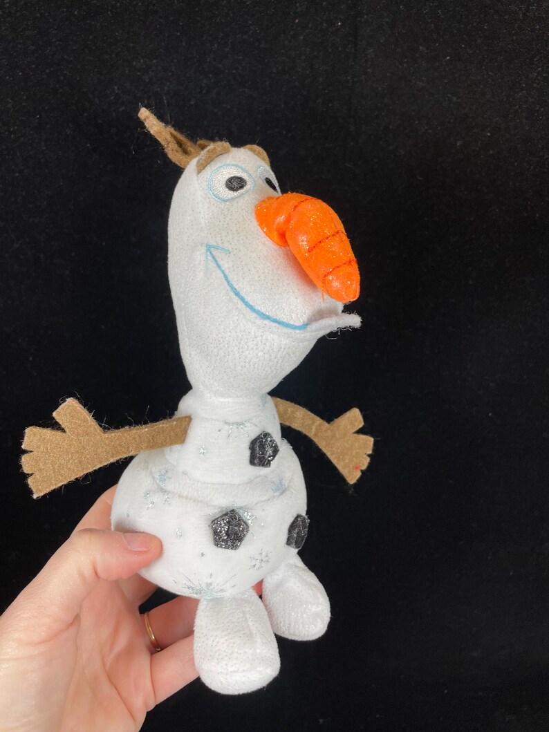 May include: A plush snowman toy with a carrot nose, brown twig arms, and a small brown twig on top of its head. The snowman has black glitter buttons and snowflake accents. The toy is white with blue embroidered eyes and a blue smile.