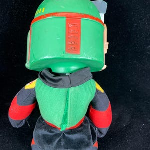May include: A soft toy of the Star Wars character Boba Fett. The toy has a green helmet with red detailing, a green and black body, and red and black arms. The design is based on the character's distinctive armour.