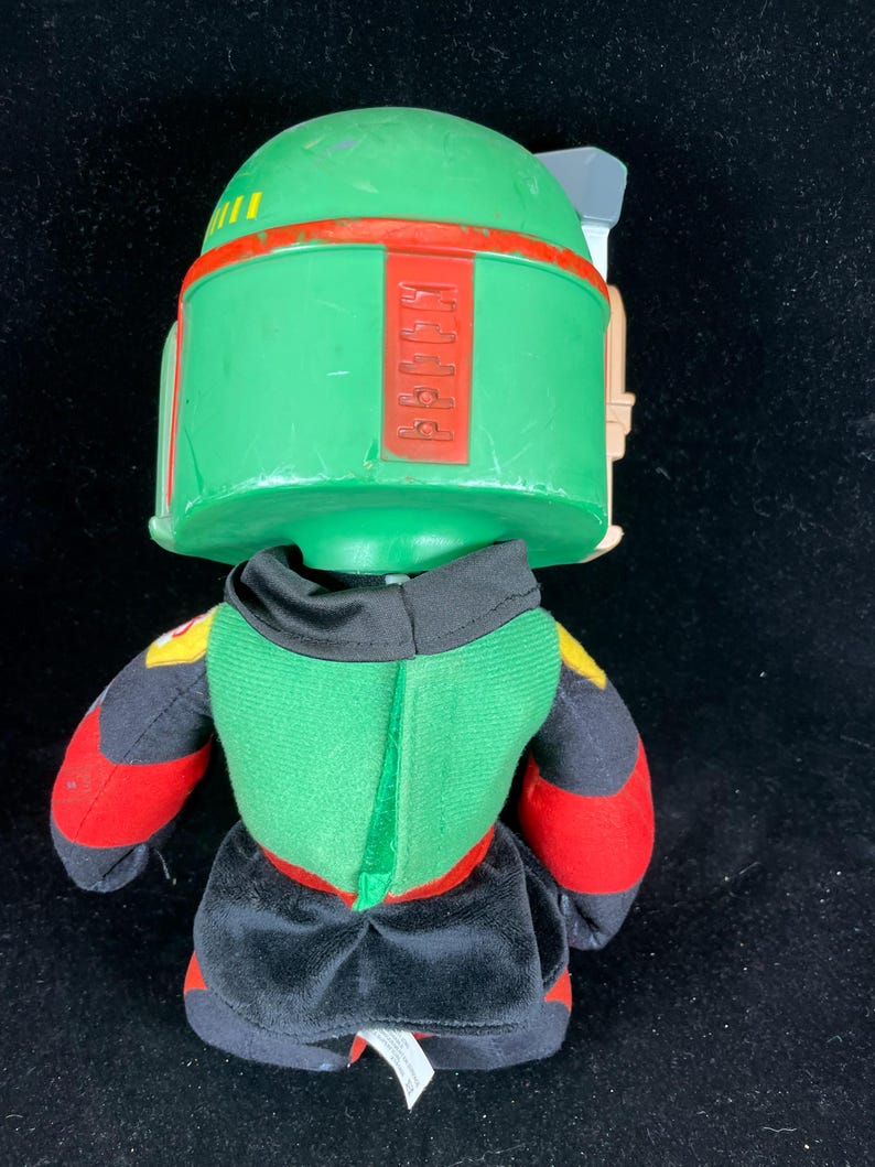 May include: A soft toy of the Star Wars character Boba Fett. The toy has a green helmet with red detailing, a green and black body, and red and black arms. The design is based on the character's distinctive armour.