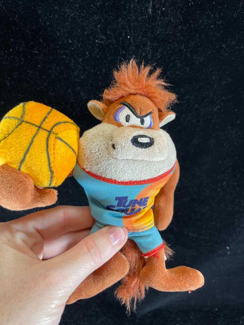 Puede incluir: Un peluche de Taz, de Space Jam, sosteniendo una pelota de baloncesto. Taz lleva una camiseta azul y naranja con las palabras "Tune Squad". El peluche tiene pelaje marr&oacute;n, pelo naranja y una expresi&oacute;n enfadada.