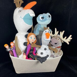 May include: A collection of Frozen-themed plush toys and figurines in a light gray basket. The characters include Olaf, Anna, Kristoff, Sven, and Bruni. The toys are set against a black background.