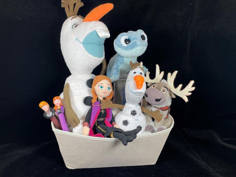 May include: A collection of Frozen-themed plush toys and figurines in a light gray basket. The characters include Olaf, Anna, Kristoff, Sven, and Bruni. The toys are set against a black background.