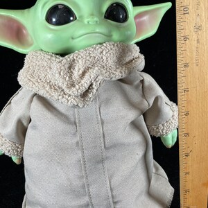 May include: A green plush toy of a character from the Star Wars franchise, known as "The Child" or "Baby Yoda", wearing a brown hooded coat. The toy is approximately 10 inches tall and is standing next to a wooden ruler.