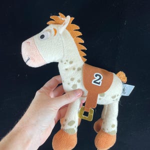May include: A plush toy horse with a cream-colored body, brown spots, and orange mane and hooves. It has a brown saddle with the number "2" on it. The toy is a character from the Toy Story franchise.