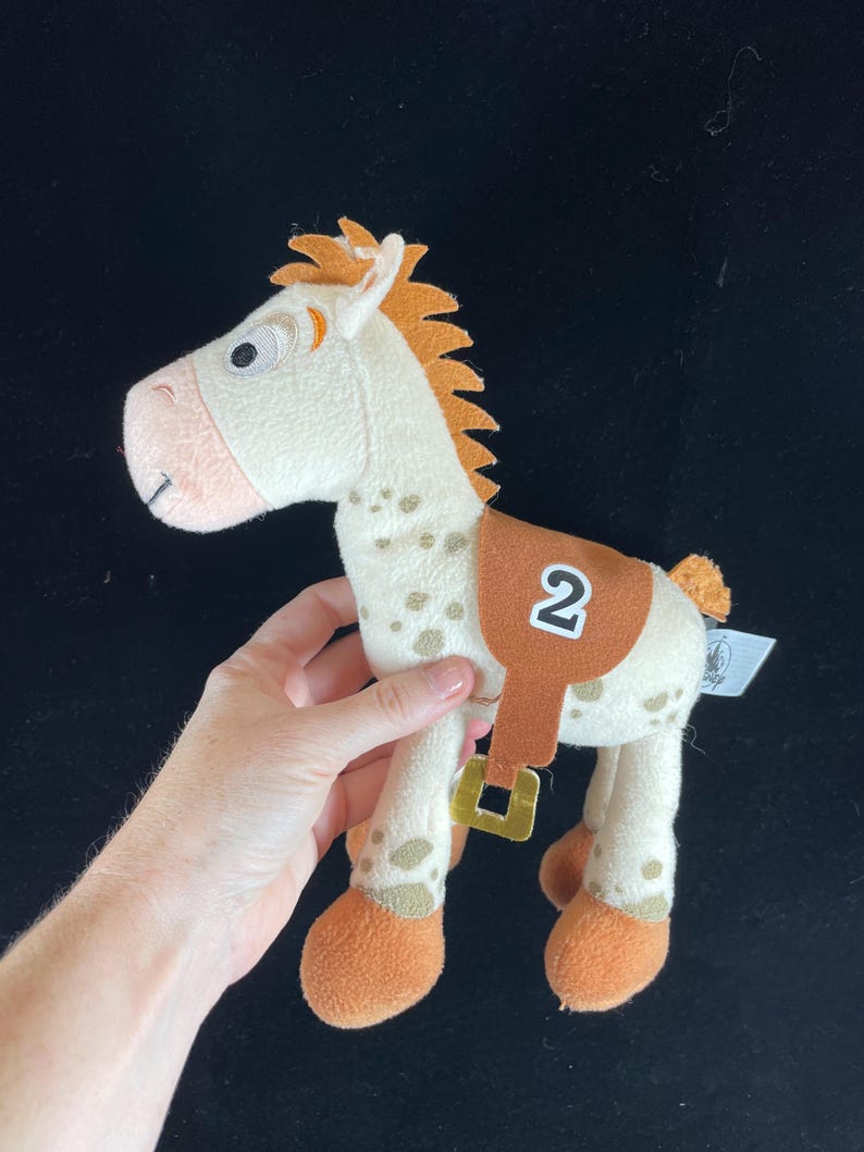 May include: A plush toy horse with a cream-colored body, brown spots, and orange mane and hooves. It has a brown saddle with the number "2" on it. The toy is a character from the Toy Story franchise.