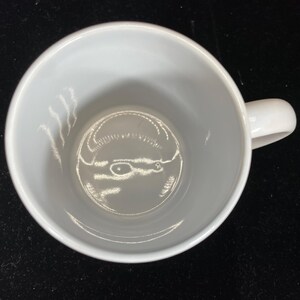 Op de afbeelding: Een witte keramische koffiemok met een gebogen oor. De mok is leeg en van bovenaf gezien, waardoor een gladde, schone binnenkant zichtbaar is. De mok staat op een donkere ondergrond, wat een contrast cre&euml;ert met de helderwitte kleur.