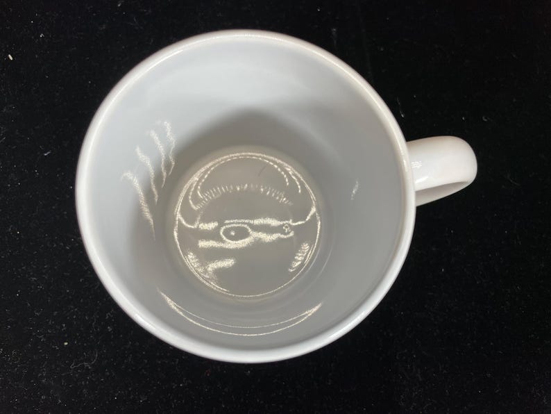 Op de afbeelding: Een witte keramische koffiemok met een gebogen oor. De mok is leeg en van bovenaf gezien, waardoor een gladde, schone binnenkant zichtbaar is. De mok staat op een donkere ondergrond, wat een contrast cre&euml;ert met de helderwitte kleur.