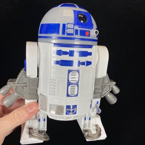 Op de afbeelding: Een wit en blauw R2-D2 speelgoedrobot met grijze accenten. Het speelgoed heeft een afgeronde kop, een rechthoekig lichaam en meerdere mechanische details. De robot is een Star Wars verzamelobject.