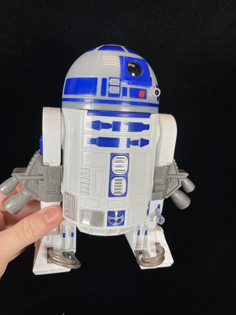 Op de afbeelding: Een wit en blauw R2-D2 speelgoedrobot met grijze accenten. Het speelgoed heeft een afgeronde kop, een rechthoekig lichaam en meerdere mechanische details. De robot is een Star Wars verzamelobject.