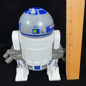 Op de afbeelding: Een wit en blauw R2-D2 speelgoedrobot met een grijze kop. Het speelgoed heeft een liniaal ernaast, dat de hoogte aangeeft. De robot heeft een gedetailleerd ontwerp met blauwe en grijze accenten en is een verzamelobject.
