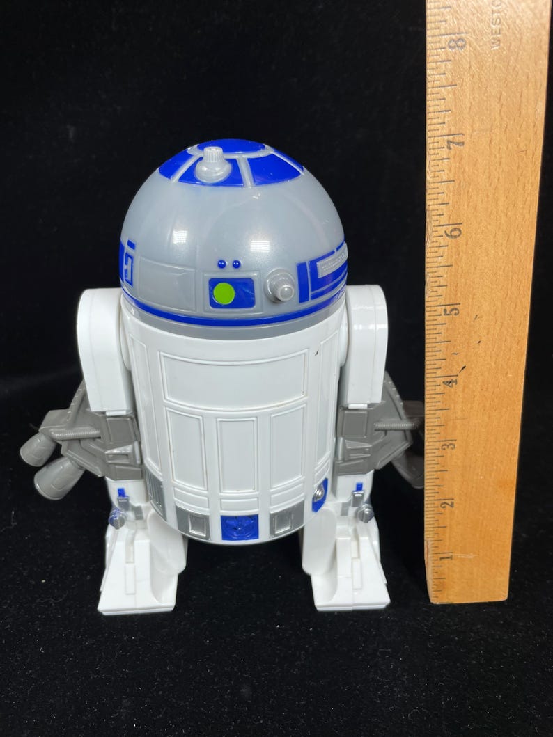 Op de afbeelding: Een wit en blauw R2-D2 speelgoedrobot met een grijze kop. Het speelgoed heeft een liniaal ernaast, dat de hoogte aangeeft. De robot heeft een gedetailleerd ontwerp met blauwe en grijze accenten en is een verzamelobject.
