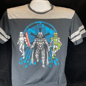 May include: Black t-shirt with a grey yoke and sleeves, featuring a pixelated graphic of Darth Vader, a Stormtrooper, and Boba Fett. The design includes a blue Imperial symbol and a red lightsaber. The shirt has grey stripes on the sleeves.