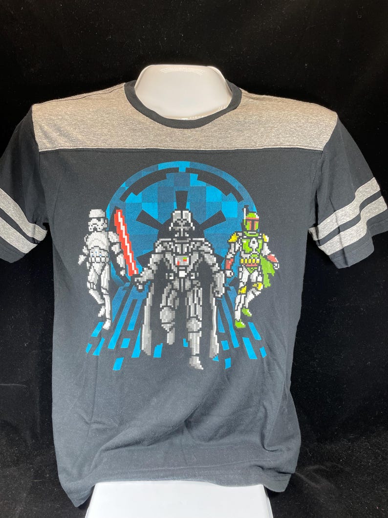 May include: Black t-shirt with a grey yoke and sleeves, featuring a pixelated graphic of Darth Vader, a Stormtrooper, and Boba Fett. The design includes a blue Imperial symbol and a red lightsaber. The shirt has grey stripes on the sleeves.
