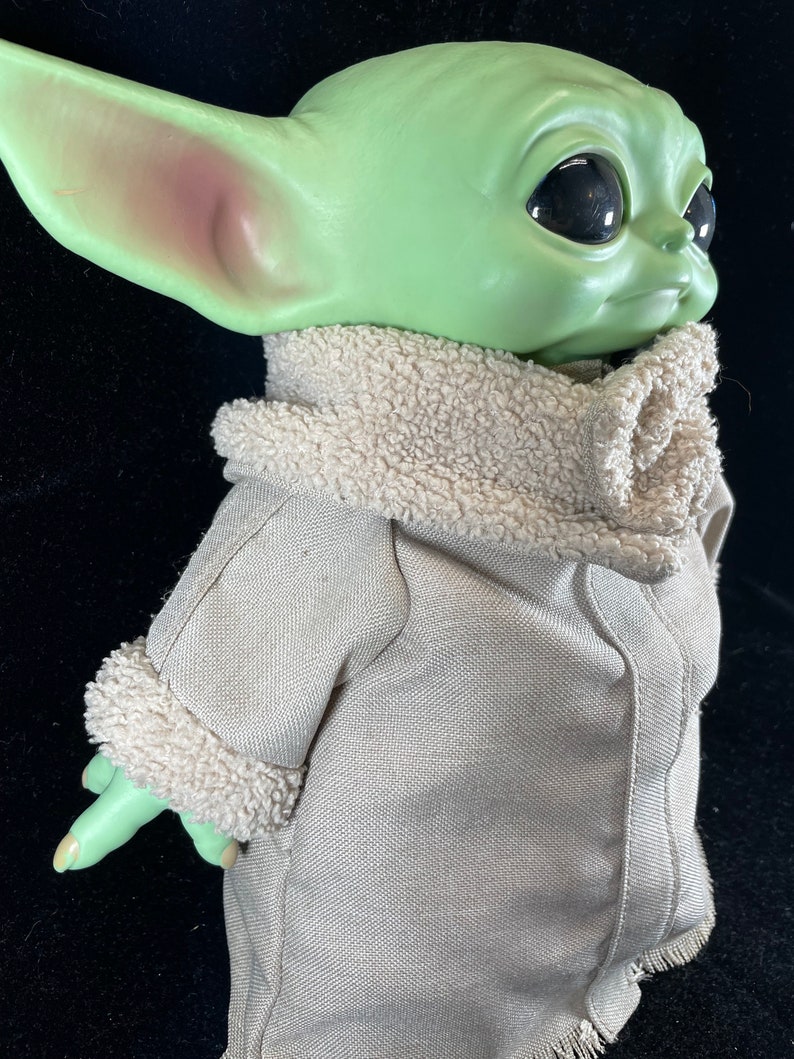 May include: A green plush toy with large eyes and a brown fur collar wearing a gray jacket. The toy is a popular character from the Star Wars franchise.