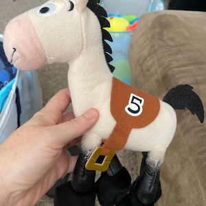 May include: A plush toy horse with a cream-colored body, black mane and tail, and a brown saddle with the number 5. The horse has black hooves and a gold-colored buckle. The toy is designed to resemble a cartoon character.