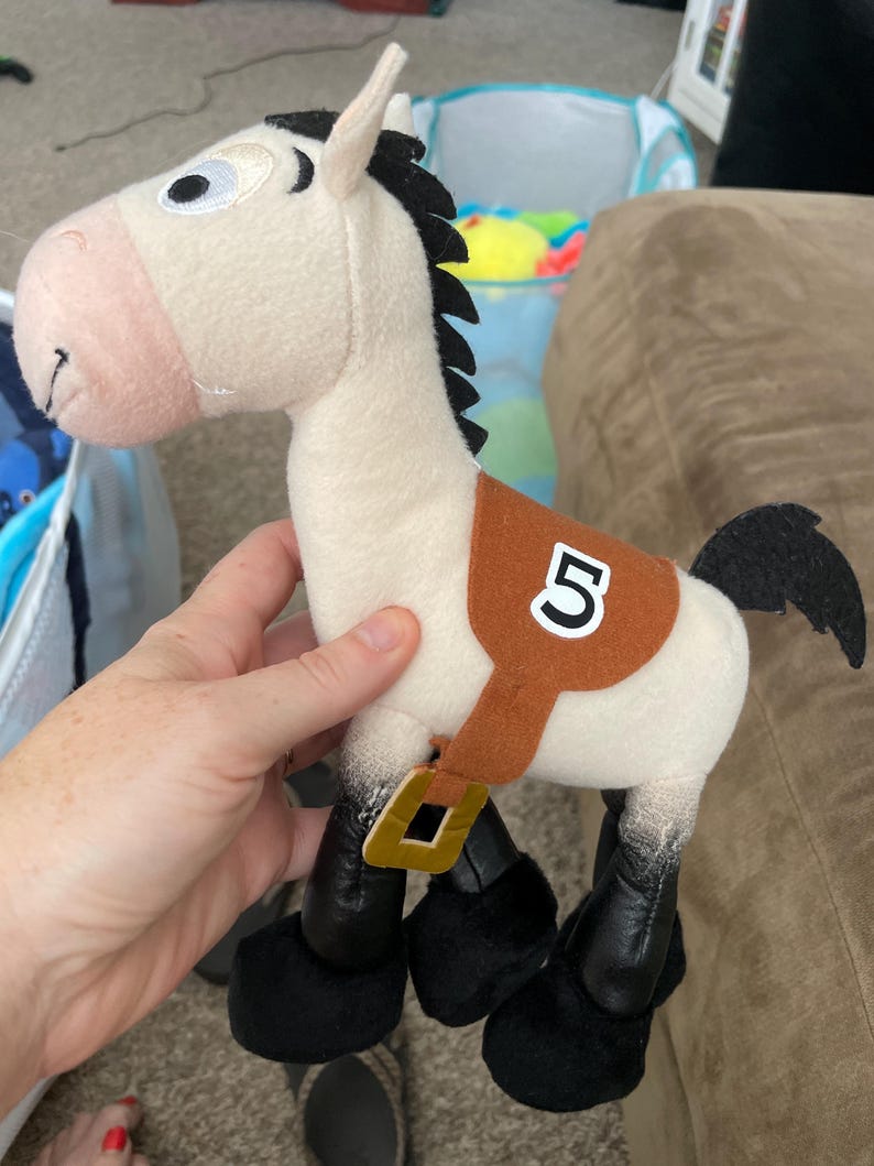 May include: A plush toy horse with a cream-colored body, black mane and tail, and a brown saddle with the number 5. The horse has black hooves and a gold-colored buckle. The toy is designed to resemble a cartoon character.