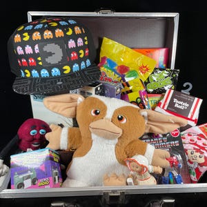 May include: A silver metal case filled with various items. A black Pac-Man themed hat sits atop the contents. Other items include a Gremlins plush, candy, a Strawberry Shortcake item, and a toy robot.