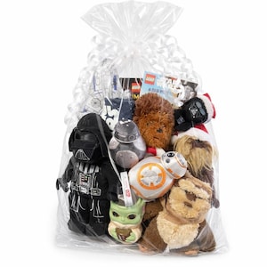 Puede incluir: Una bolsa de regalo de plástico transparente llena de artículos con temática de Star Wars, incluyendo un peluche negro de Darth Vader, un peluche marrón de Chewbacca, un droide BB-8 redondo, una figura verde de Grogu y una caja LEGO Star Wars, con lazo blanco.