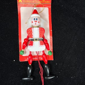 May include: A wooden Christmas ornament in the shape of a Santa Claus figure. The figure is red and white with black boots and green hands. The ornament is attached to a red cardboard backing with the text "Visions of Sugar Plums Collection".