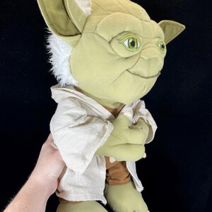 Op de afbeelding: Een pluche Yoda-pop uit Star Wars. De pop heeft grote groene oren, een lichtgroen gezicht en een bruine jurk. De ogen van de pop zijn groen en ze heeft wit haar rond de oren. De pop houdt haar handen bij elkaar.