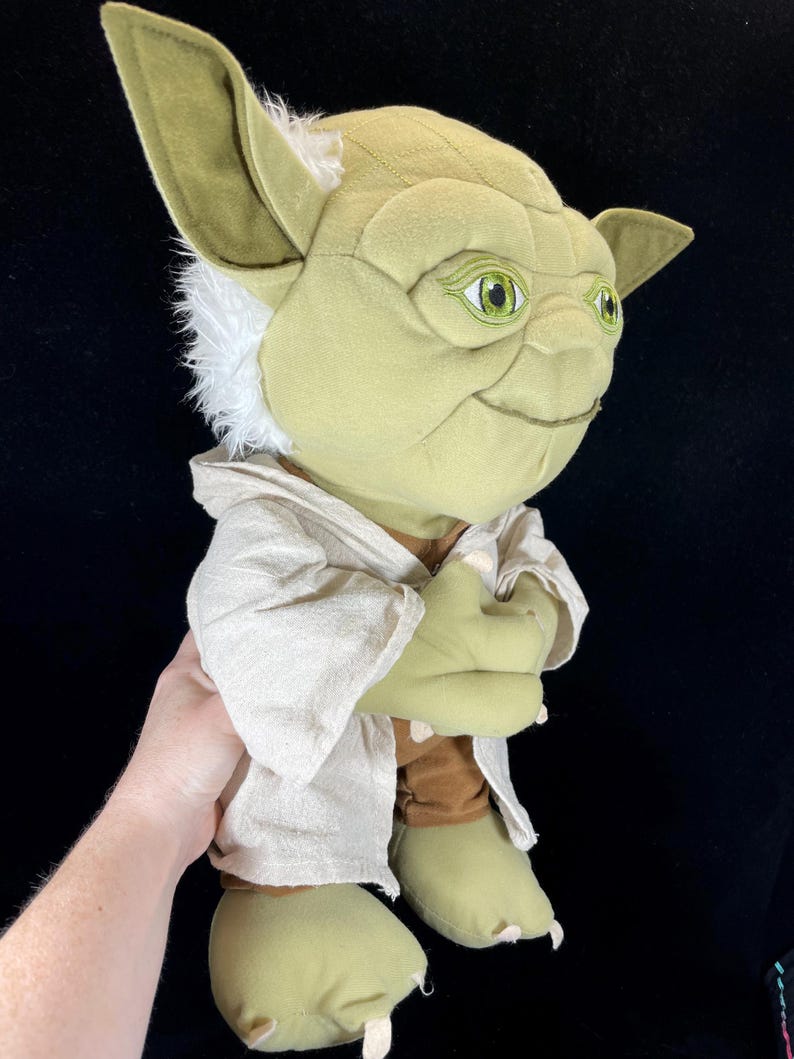 Op de afbeelding: Een pluche Yoda-pop uit Star Wars. De pop heeft grote groene oren, een lichtgroen gezicht en een bruine jurk. De ogen van de pop zijn groen en ze heeft wit haar rond de oren. De pop houdt haar handen bij elkaar.